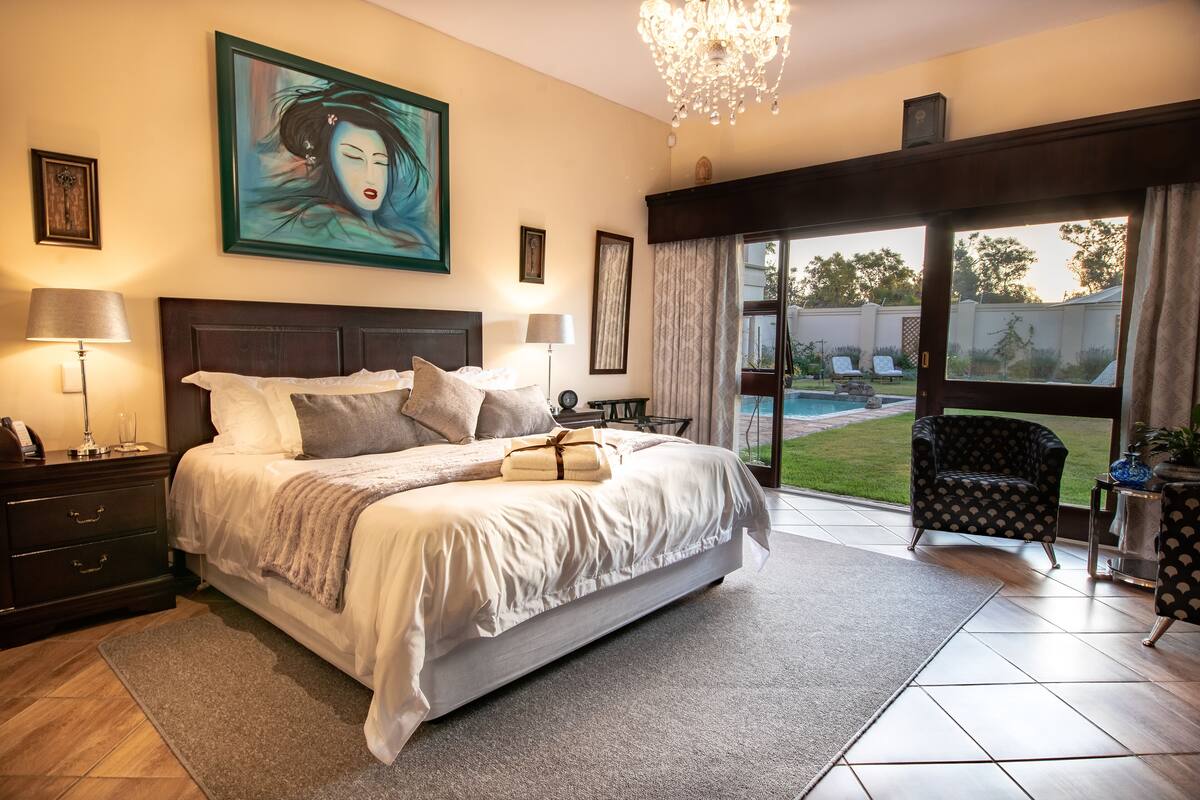 A bedroom with a large bed, a chandelier, a painting, and a view of the outdoors.