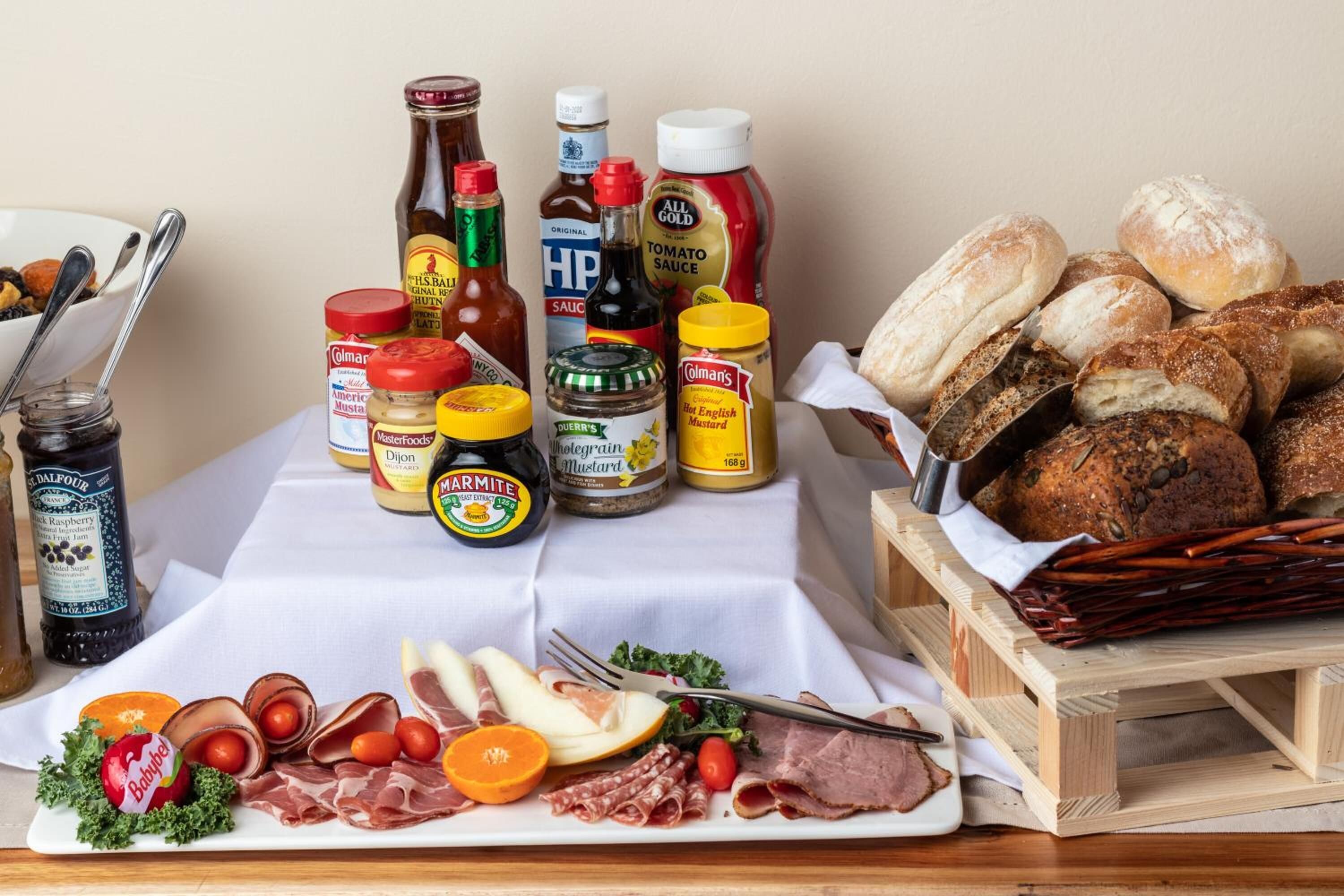 Uma mesa com diversos alimentos, incluindo carnes, queijos e acompanhamentos, além de uma cesta de pães.
