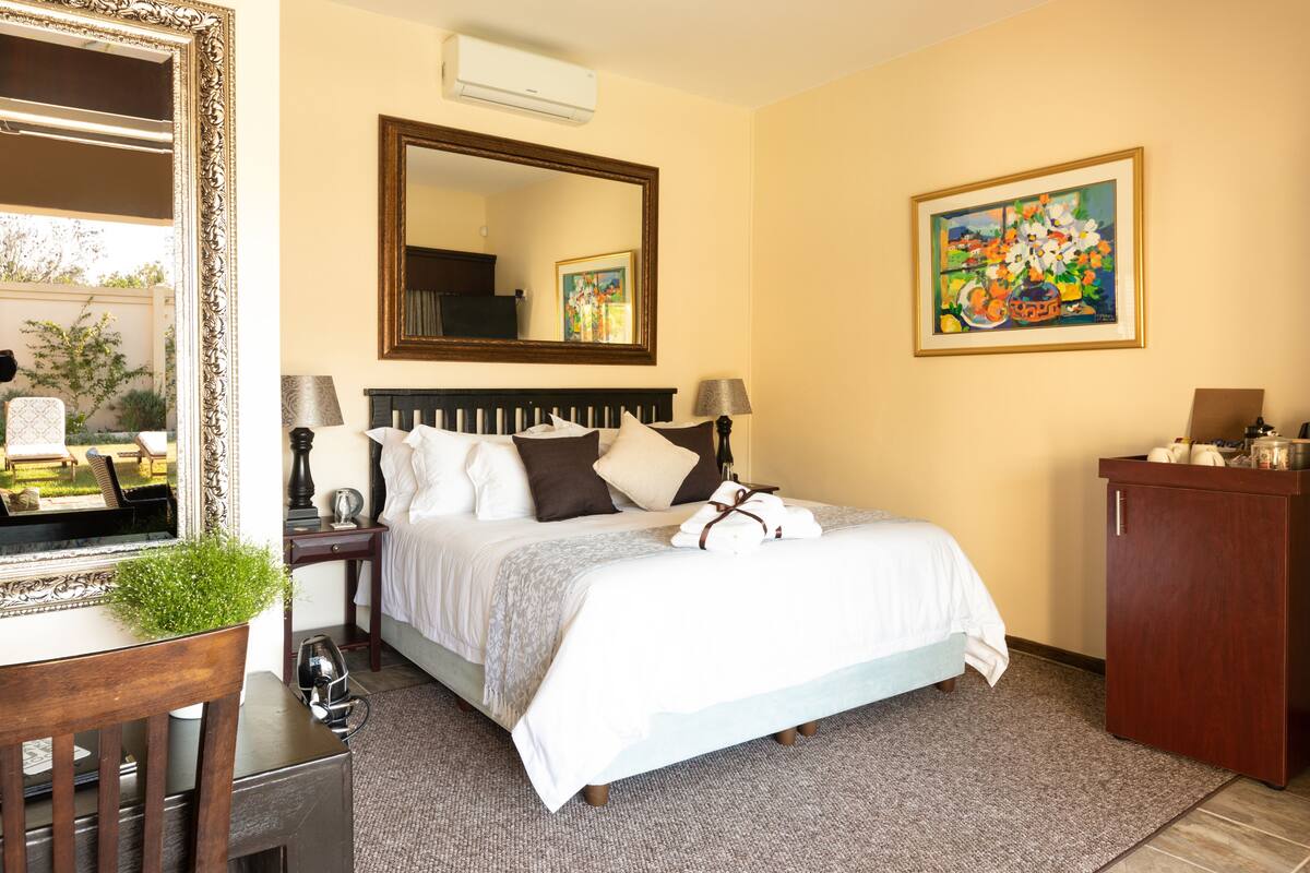 A hotel room with a large bed, a wooden headboard, a bedside table with a lamp, a dresser, a mirror, and a painting on the wall.