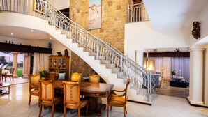 A spacious living area with a large wooden dining table, comfortable seating, a staircase, and a view of the outdoors. - Amery House (Gqeberha (Port Elizabeth))