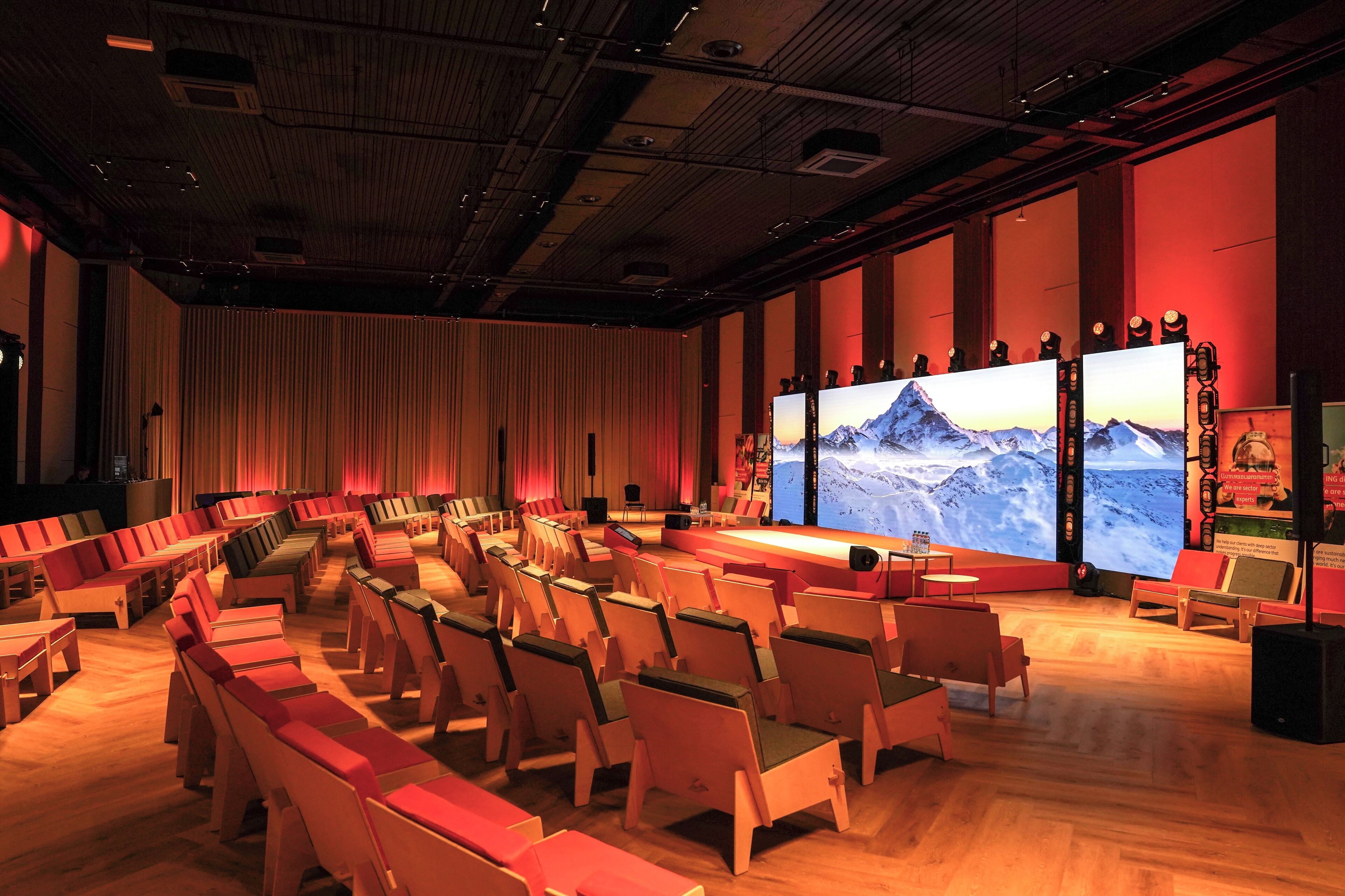 Ein Theater mit Reihen aus Holzstühlen und einer großen Leinwand, die eine Berglandschaft zeigt.