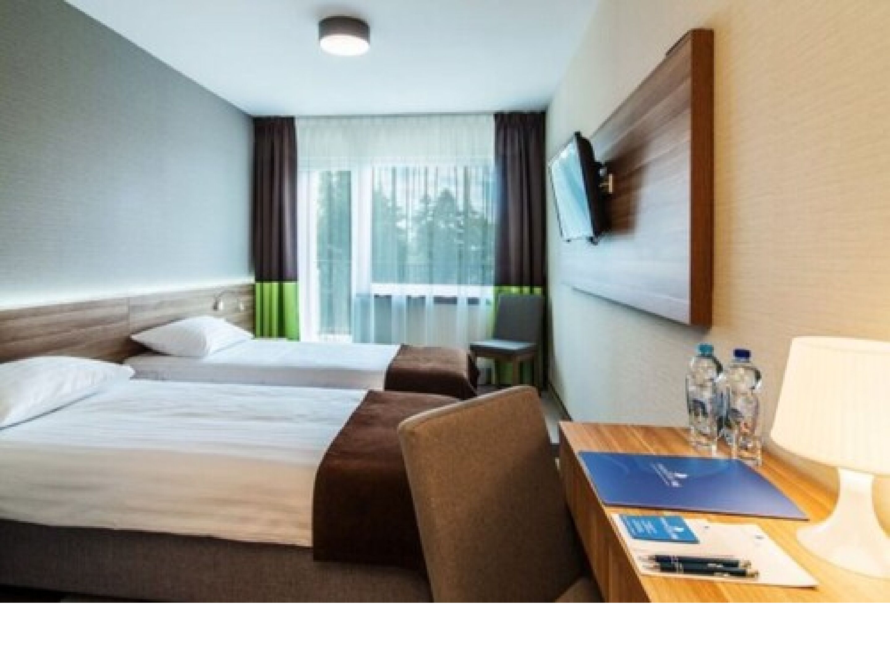 A hotel room with two beds, a desk with a chair, a TV mounted on the wall, and a window with curtains.