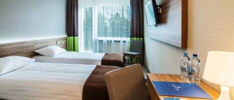 Habitación de hotel con dos camas, un escritorio con silla, un televisor montado en la pared y una ventana con cortinas.