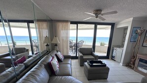 A spacious living room with a large sofa, armchairs, and a dining area. A ceiling fan is visible, and the room has large windows offering a view of the beach. - Gulf Front Condo with Stunning Views! (Pensacola)