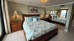 A spacious bedroom with a large bed, a chandelier, a painting, and a view of the sea through a window. - Gulf Front Condo with Stunning Views! (Pensacola)