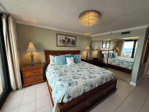 A spacious bedroom with a large bed, a chandelier, a painting, and a view of the sea through a window.