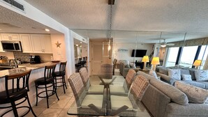 A modern living space with a glass-top dining table surrounded by wicker chairs, a grey sofa, and a kitchen area with white cabinets and a microwave.