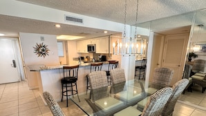 A dining area with a glass-top table and wicker chairs, a kitchen in the background, and a ceiling vent.
