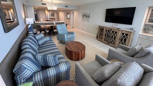 A living room with a sofa, armchairs, a coffee table, a TV, and a kitchen area in the background. - Eden West 905 Completely updated Gulf Front Condo with Stunning Views! (Pensacola)
