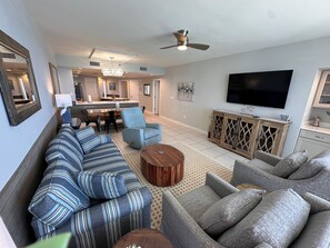 A living room with a sofa, armchairs, a coffee table, a TV, and a kitchen area in the background.