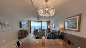 A dining area with a wooden table, chairs, a chandelier, a sofa, and a view of the ocean. - Eden West 905 Completely updated Gulf Front Condo with Stunning Views! (Pensacola)