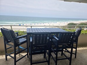 Une table et des chaises en bois sur un balcon donnant sur une plage de sable fin, avec l’océan et ses vagues en toile de fond.