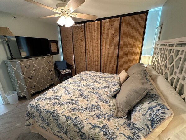 Un dormitorio con cama, televisor en una cómoda, ventilador de techo y ventana con persianas.