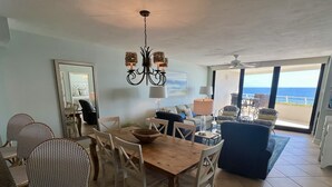 Een eetruimte met een houten tafel en witte stoelen, een kroonluchter, en uitzicht op de oceaan door grote ramen.