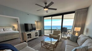 A spacious living room with a large sofa, a coffee table, and a TV on a cabinet. There is a balcony with a view of the ocean. - Updated W 700 1 Bedroom 2 Bath Gulf Front Condo with Stunning Views! (Pensacola)