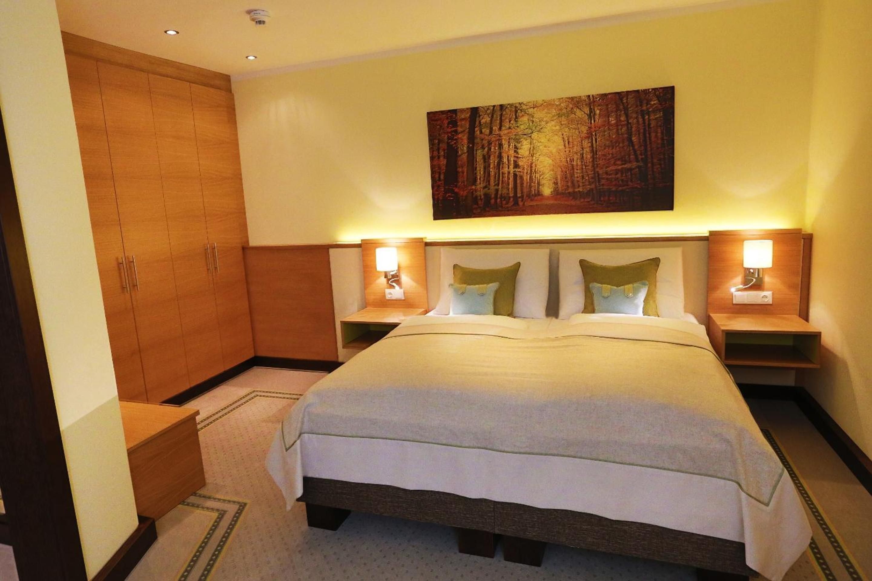 A hotel room with a large bed, two bedside tables with lamps, a wardrobe, and a painting on the wall.