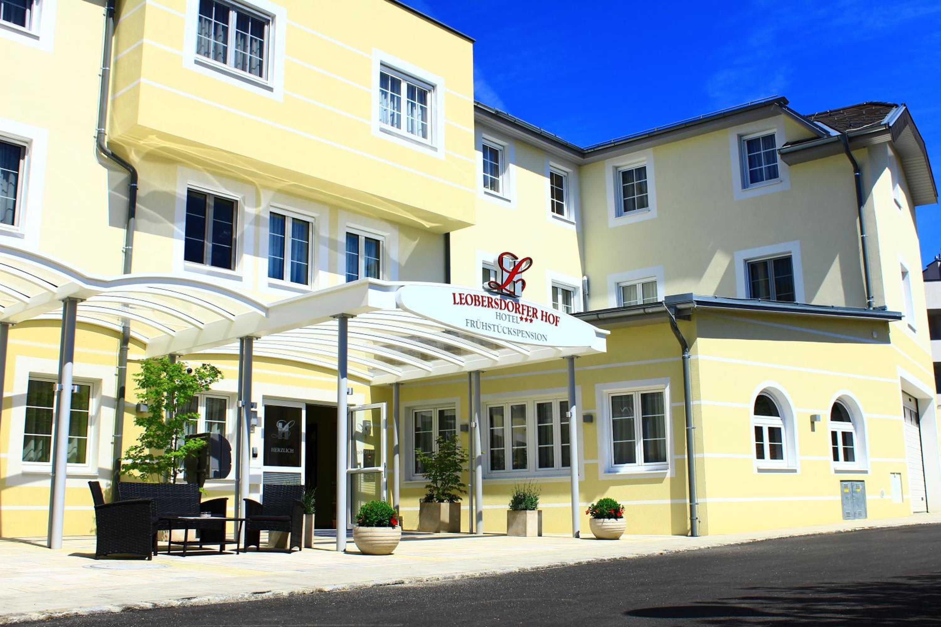 Ein Hotel mit gelber Fassade, überdachter Eingangspartei, Außenbestuhlung und Pflanzkübeln.