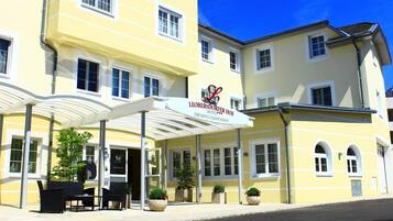 Ein Hotel mit gelber Fassade, überdachter Eingangspartei, Außenbestuhlung und Pflanzkübeln.