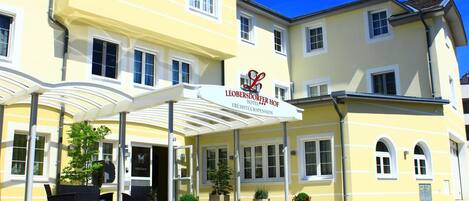 A hotel with a yellow facade, a covered entrance, outdoor seating, and potted plants.