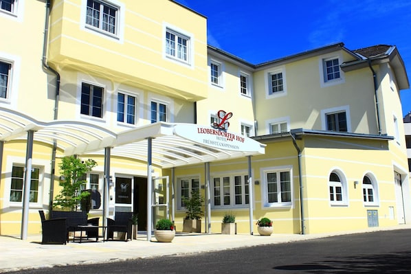 Um hotel com fachada amarela, entrada coberta, área de estar externa e vasos de plantas.