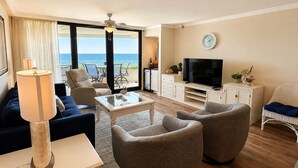 Ein Wohnzimmer mit einer Couch, Sesseln, einem Couchtisch, einem Fernseher und einem Balkon mit Meerblick.