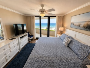 A hotel room with a bed, a TV, a ceiling fan, a balcony with a view of the ocean, and a painting on the wall.
