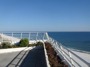 Uma passarela costeira com corrimão branco, céu azul límpido e o oceano ao fundo.
