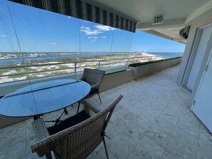 Varanda com mesa de vidro e cadeiras de vime, com vista para uma praia e um horizonte urbano distante.
