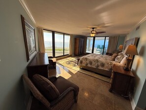 Quarto com cama, poltrona, escrivaninha, ventoinha no teto e vista para o oceano através de amplas janelas.