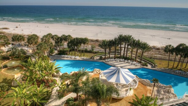 Een resort met een groot zwembad, palmbomen en uitzicht op het strand.
