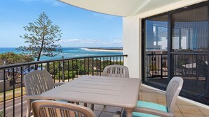 En balkon med et bord og stole, der vender ud mod et kystlandskab med et træ og en bygning i baggrunden.
