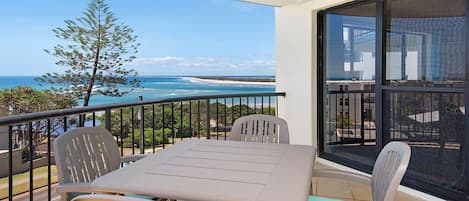 A balcony with a table and chairs, overlooking a coastal view with a tree and a building in the background.