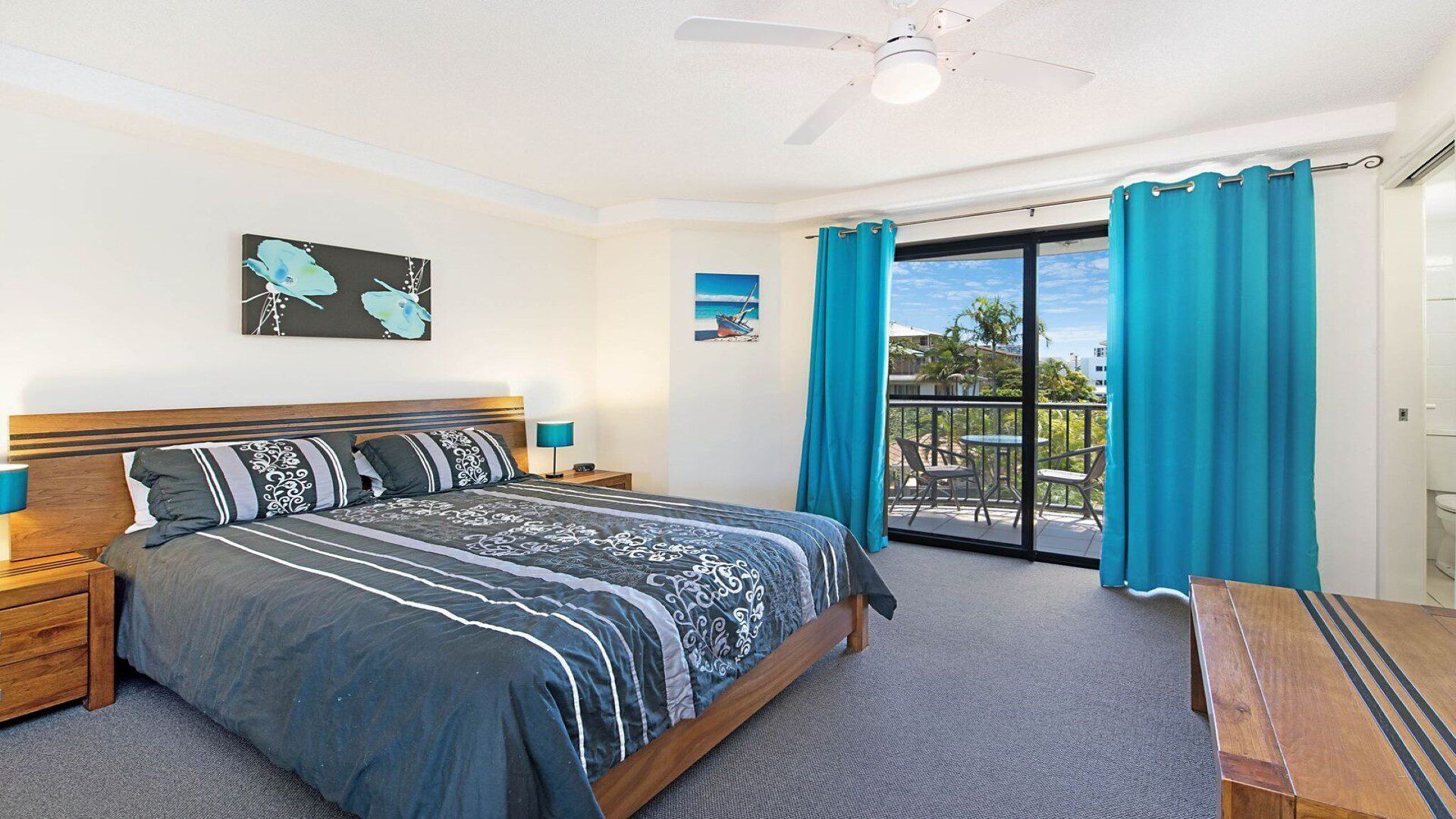 A bedroom with a bed, a nightstand, a desk, a ceiling fan, a balcony with a view, and a painting on the wall.