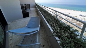 A balcony with a view of a beach, a lounge chair, and a small table with chairs.