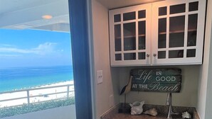 A compact kitchen with a view of the beach, a sign saying "Life is Good at the Beach", and a small refrigerator.
