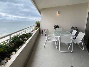 A balcony with a glass table and chairs, a fan, and a view of the sea.
