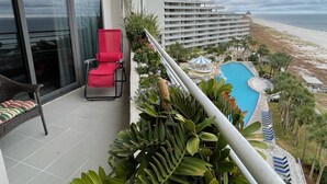 A balcony with a red chair, a pool, and a view of the beach and hotel buildings. - Gulf Front Condo with Stunning Views! (Pensacola)