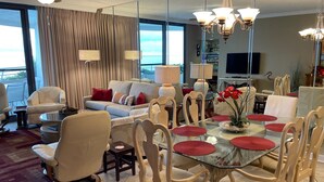 A modern living room with a glass dining table, comfortable seating, and a large window offering a view. - Gulf Front Condo with Stunning Views! (Pensacola)