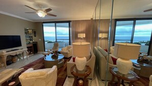 A spacious living room with a large flat-screen TV, comfortable seating, and a view of the ocean through large windows. - Gulf Front Condo with Stunning Views! (Pensacola)