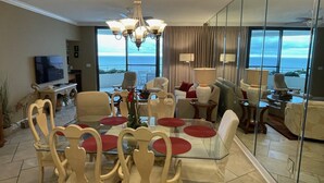 A modern living room with a glass dining table, comfortable seating, a chandelier, and a large window offering a view of the ocean. - Gulf Front Condo with Stunning Views! (Pensacola)