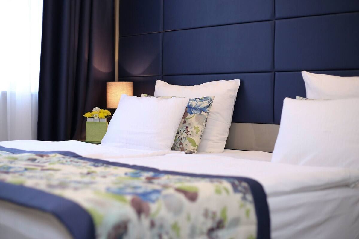 A neatly made bed with white linens and floral-patterned pillows, a dark headboard, and a bedside table with a lamp and a vase of yellow flowers.
