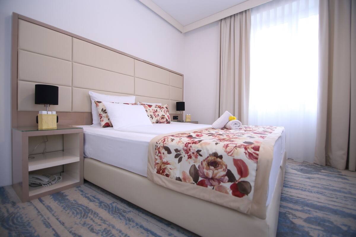 A hotel room with a large bed, a bedside table, a nightstand, a lamp, and a window with curtains.