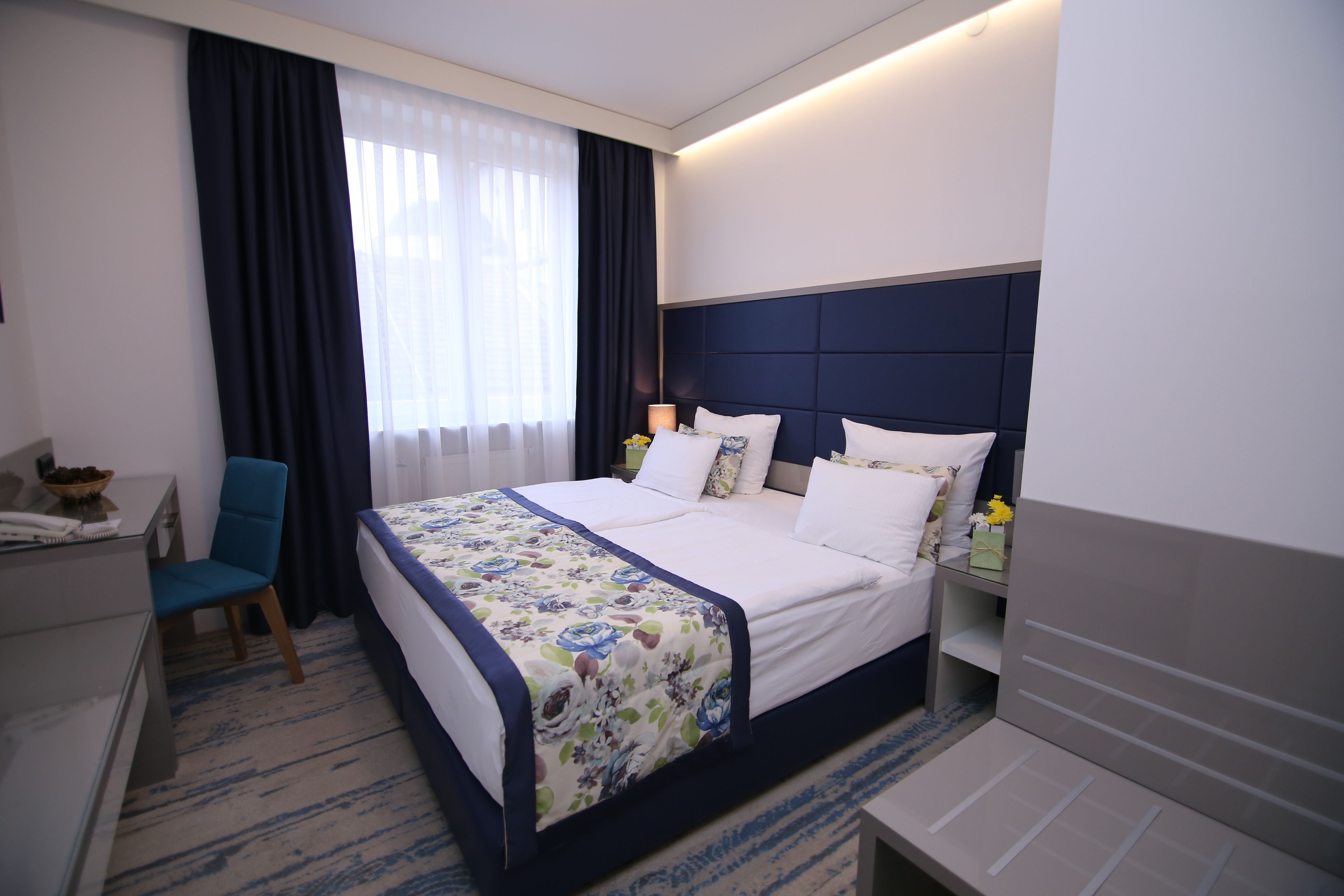 A hotel room with a large bed, a desk, a chair, and a nightstand with a lamp.
