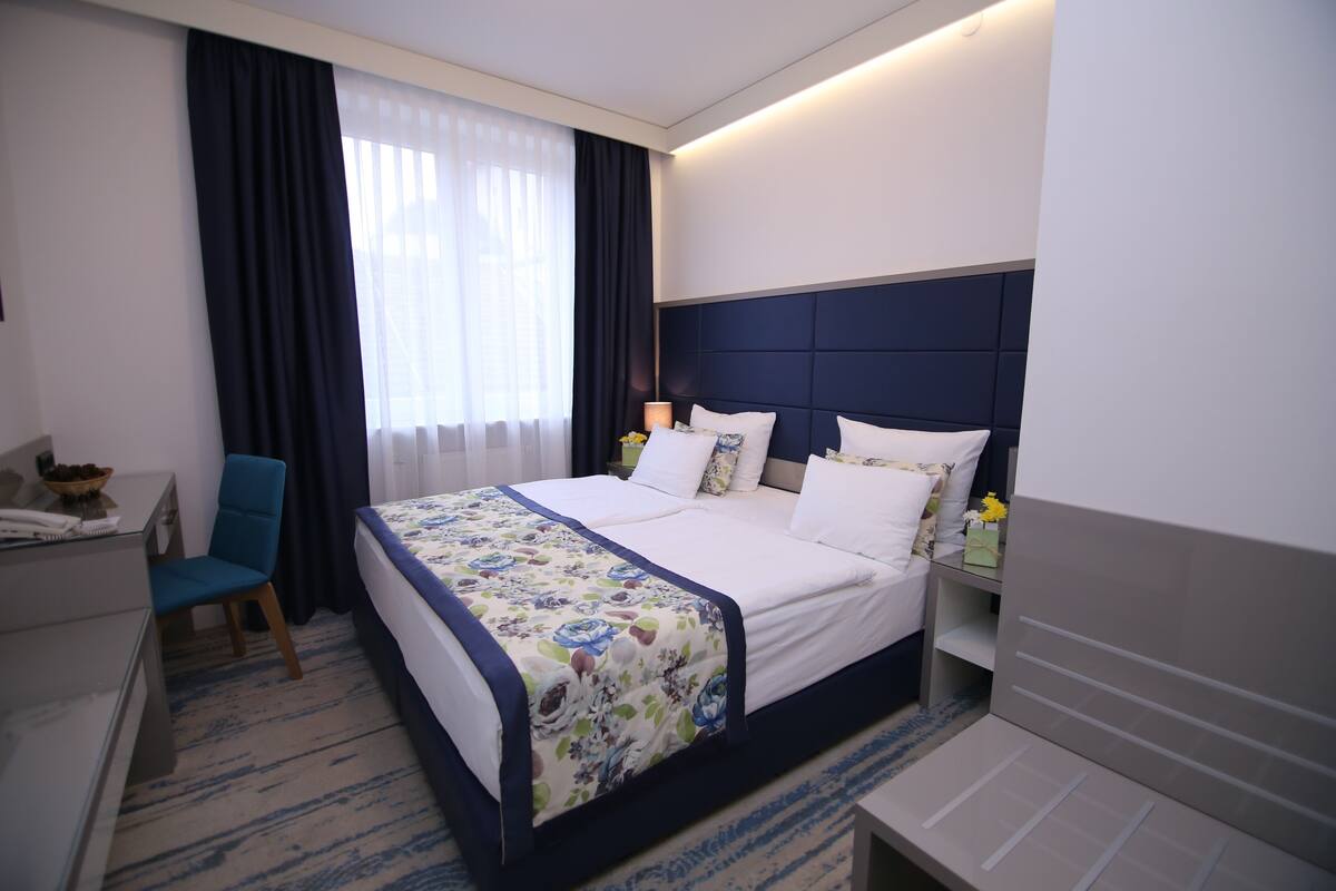 A hotel room with a large bed, a desk, a chair, and a nightstand with a lamp.