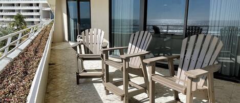A balcony with three wooden Adirondack chairs and a glass table, overlooking a body of water.