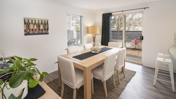A dining area with a wooden table, white chairs, a black placemat, and a painting of wine bottles on the wall. - @WineNwaves Margaret River (Margaret River)