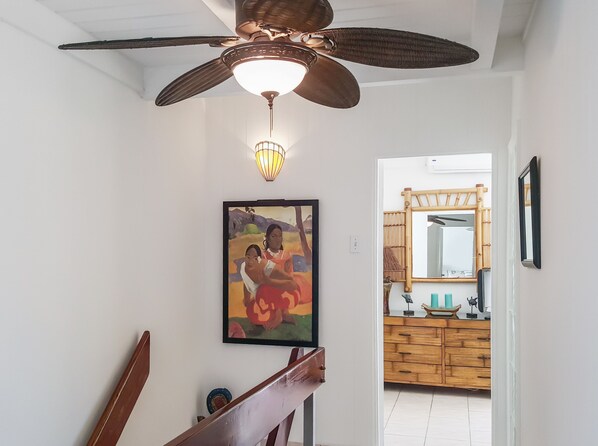 Una habitación con ventilador de techo, lámpara de pared, un cuadro enmarco y un mueble de madera con espejo.