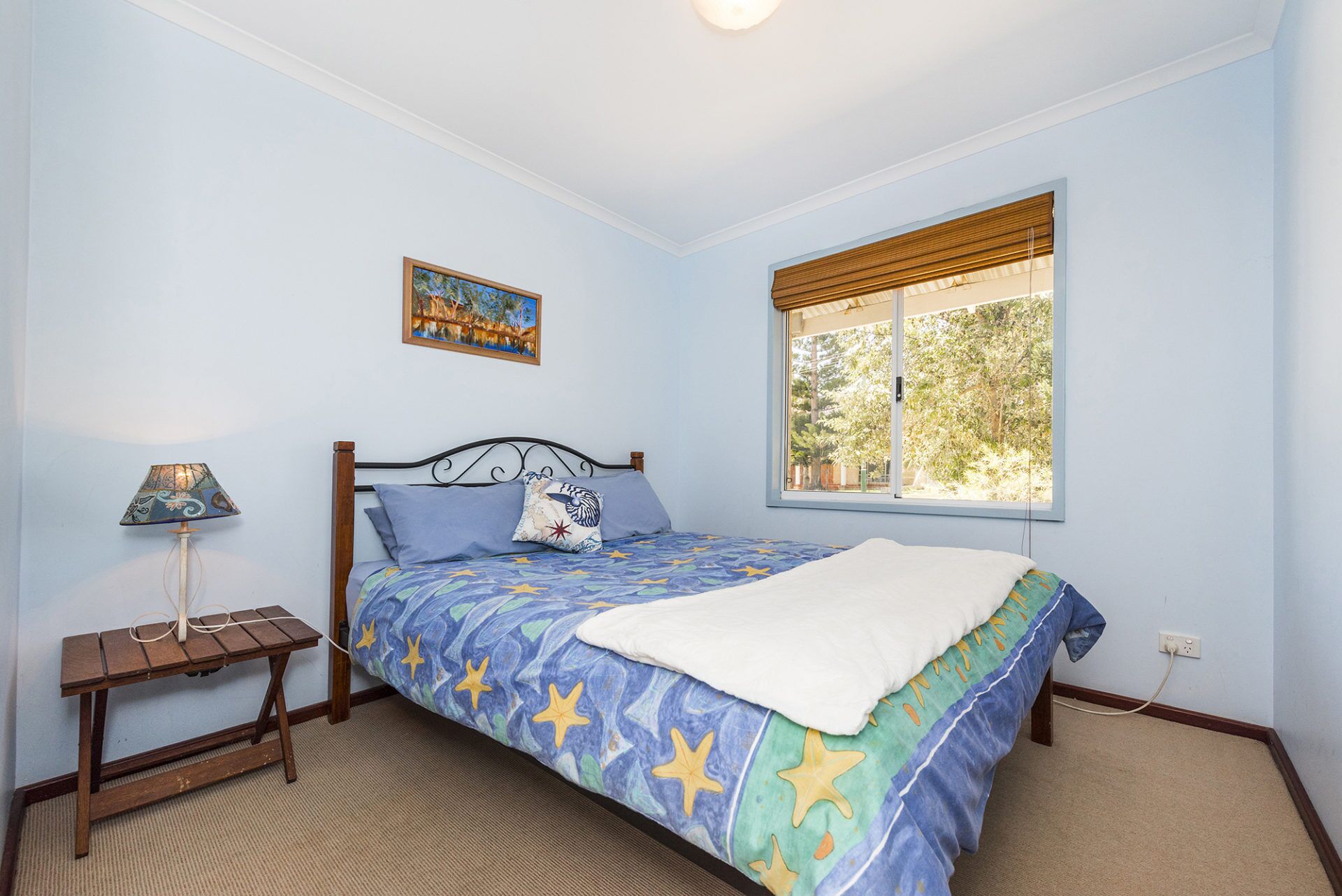 A bedroom with a bed, a nightstand, a lamp, a window with blinds, and a painting on the wall.
