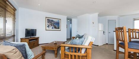Een woonkamer met een bank, een houten salontafel en een televisie op een houten dressoir. Er is een deur die leidt naar een andere kamer en een raam met jaloezieën.