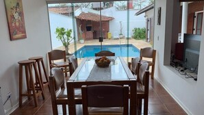 A dining area with a glass table and wooden chairs, a pool in the background, and a TV mounted on the wall. - 4 suites, air conditioning, TV, internet, swimming pool, 150 meters from the beach! Pets allowed! (Guarujá)